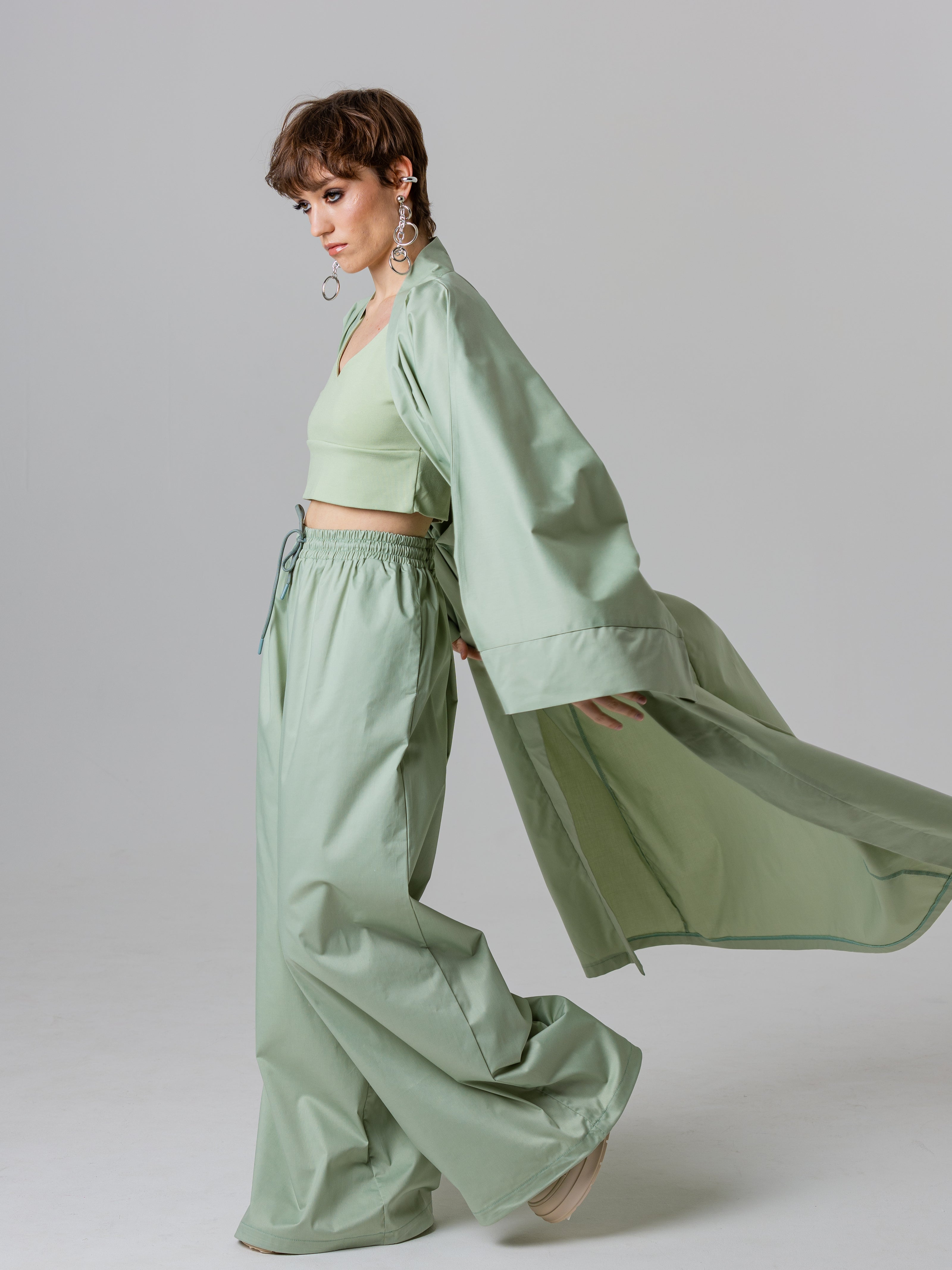 Woman wearing a light green outfit with a flowing jacket on a plain background